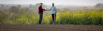 Two satisfied farmers with laptop shaking hands in yellow rapeseed field.