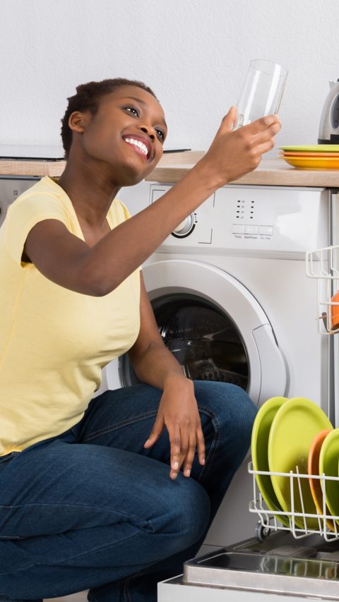 Happy African Woman Looking At Clean Glass Crouching Near Dish Washer