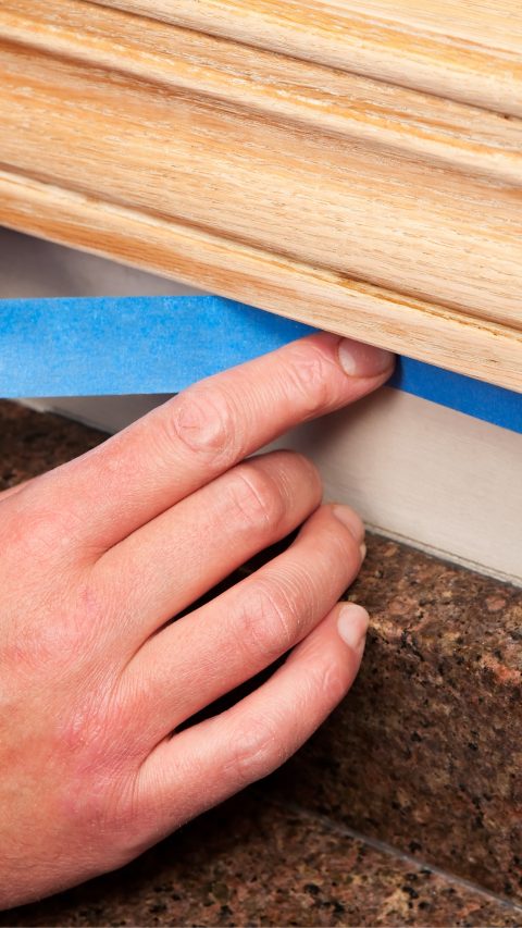 A painter is applying blue masking tape to protect an area of wall below oak window trim which is being refinished. The wall repainted after staining the trim, below the hand is a granite countertop.