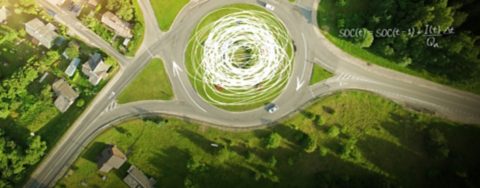 Luftaufnahme eines Kreisverkehrs mit vier Ausfahrten, umgeben von Bäumen und grünen Feldern – wissenschaftliche Gleichungen und Skizzen, die den Verkehrsfluss hervorheben, werden auf dem Bild überlagert. 