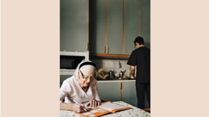 Older woman sitting at a table doing sudokus while a man is washing dishes in the background.