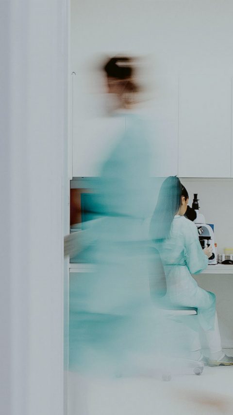 Blick in ein Schwesternzimmer einer Krankenstation. Eine Krankenschwester sitzt vor dem Computer und eine weitere Krankenschwester läuft gerade am Zimmer vorbei.