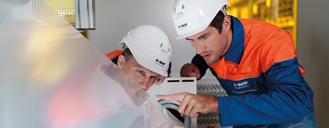 Two employees in hard hats examine a sheet of material.