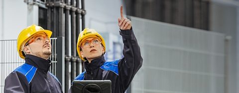 A man and woman in safety goggles and hard hats at a manufacturing site. The woman is pointing at something above them and holding a tablet device. 